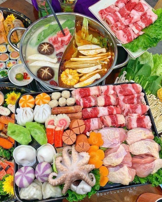 Hot pot with dual broth sections surrounded by trays of sliced meat, vegetables, mushrooms, and seafood ingredients