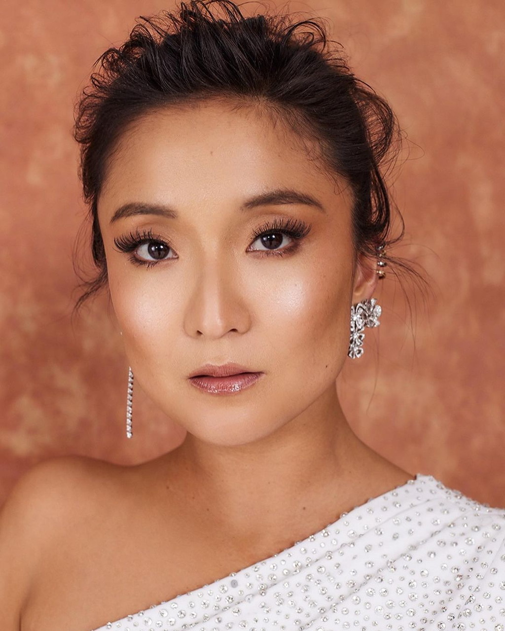 Ashley Park in a white one-shoulder outfit with sparkling earrings against a peach background.