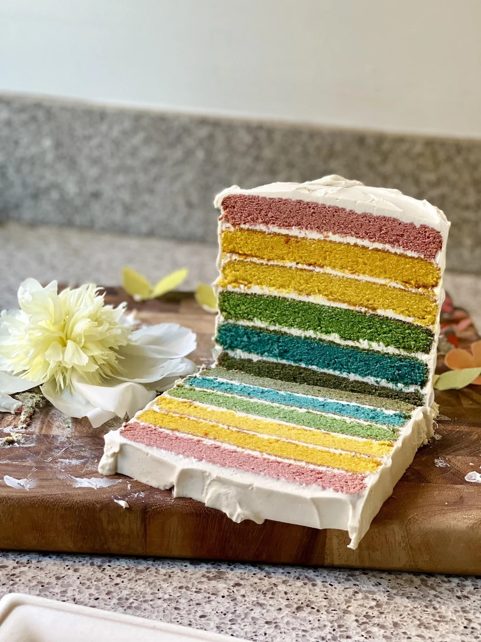 Slice of rainbow layer cake with white frosting on a wooden board next to a white flower