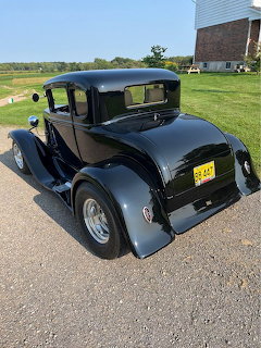 Black 1931 Ford Model A hot rod coupe parked on grass near a barn on sunny day
