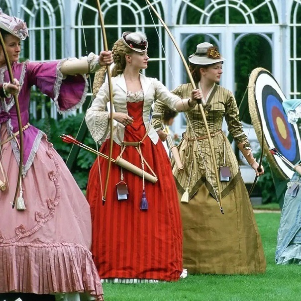 Three women in Victorian-era dresses holding bows and arrows beside an archery target outdoors