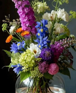 A close-up of a vibrant, colorful floral bouquet in a clear glass vase, featuring purple, blue, green, white, and orange blossoms.