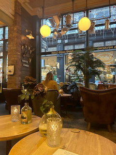 Interior of a cozy café with round wooden tables, pendant globe lights, plants, and customers by the window