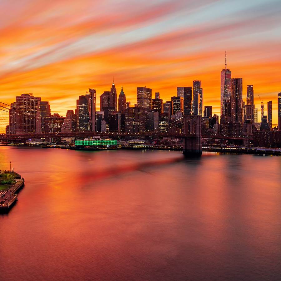 New York City skyline at sunset with orange clouds, lit skyscrapers, bridge, and water reflections