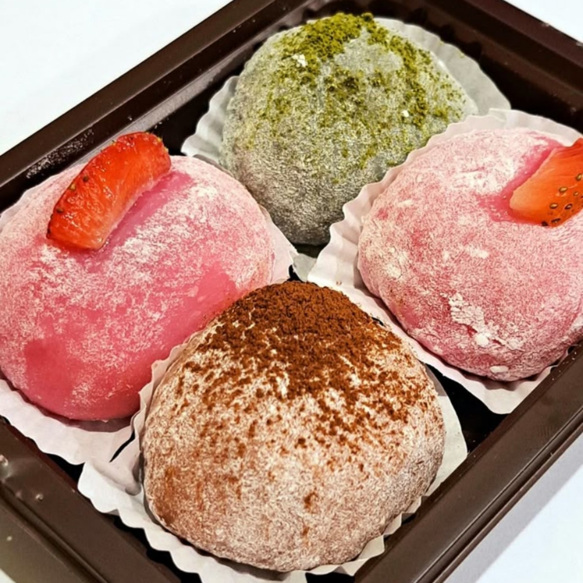 Four round mochi desserts in pink, green, and brown colors with powder coating, arranged in a box