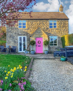 Stone cottage with pink door, garden flowers, and gravel driveway under blue sky