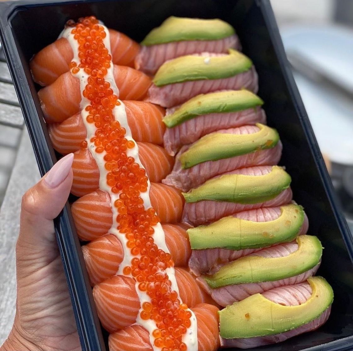 Hand holding a black tray of salmon sushi with avocado slices and orange roe