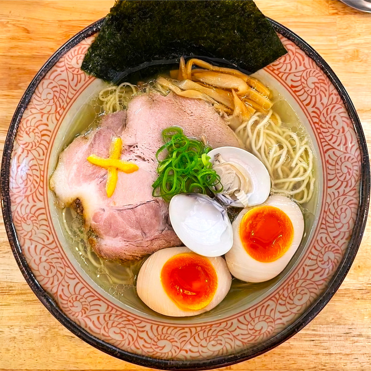 Top-down ramen bowl with noodles, pork, eggs, clams, seaweed, and green onions on a wooden table