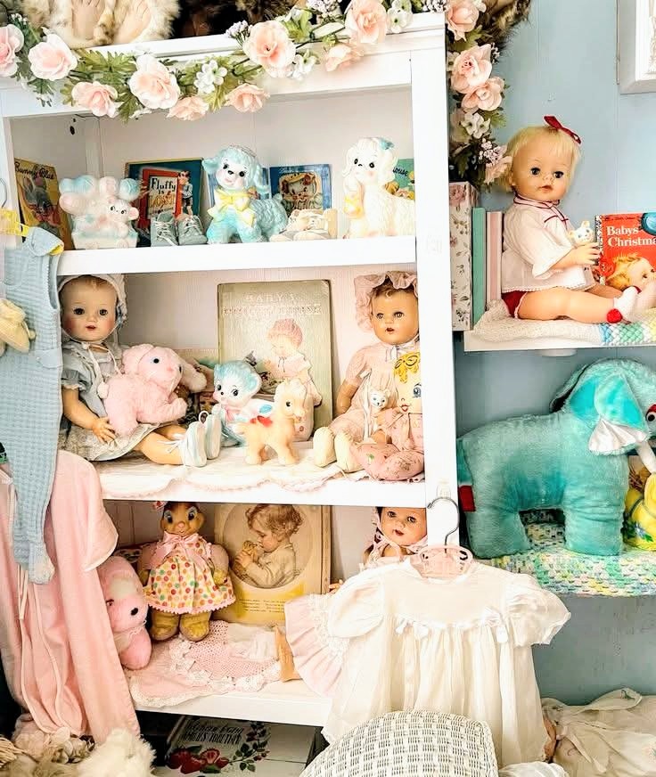White shelving unit displaying collection of vintage baby dolls, plush toys, and nursery items with floral garland