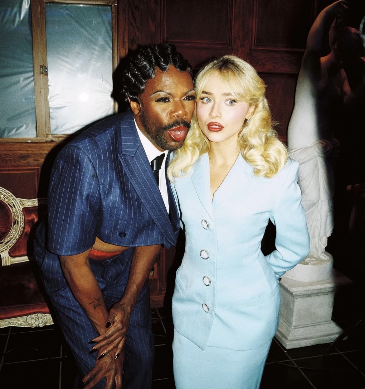 Blonde woman in light blue dress and man in navy pinstripe suit posing together in vintage interior
