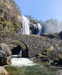 Tall waterfall flowing behind an old stone arch bridge surrounded by rocky cliffs and green vegetation