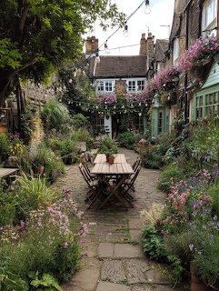 Stone courtyard garden with flowers, greenery, and a wooden table between rustic cottage buildings